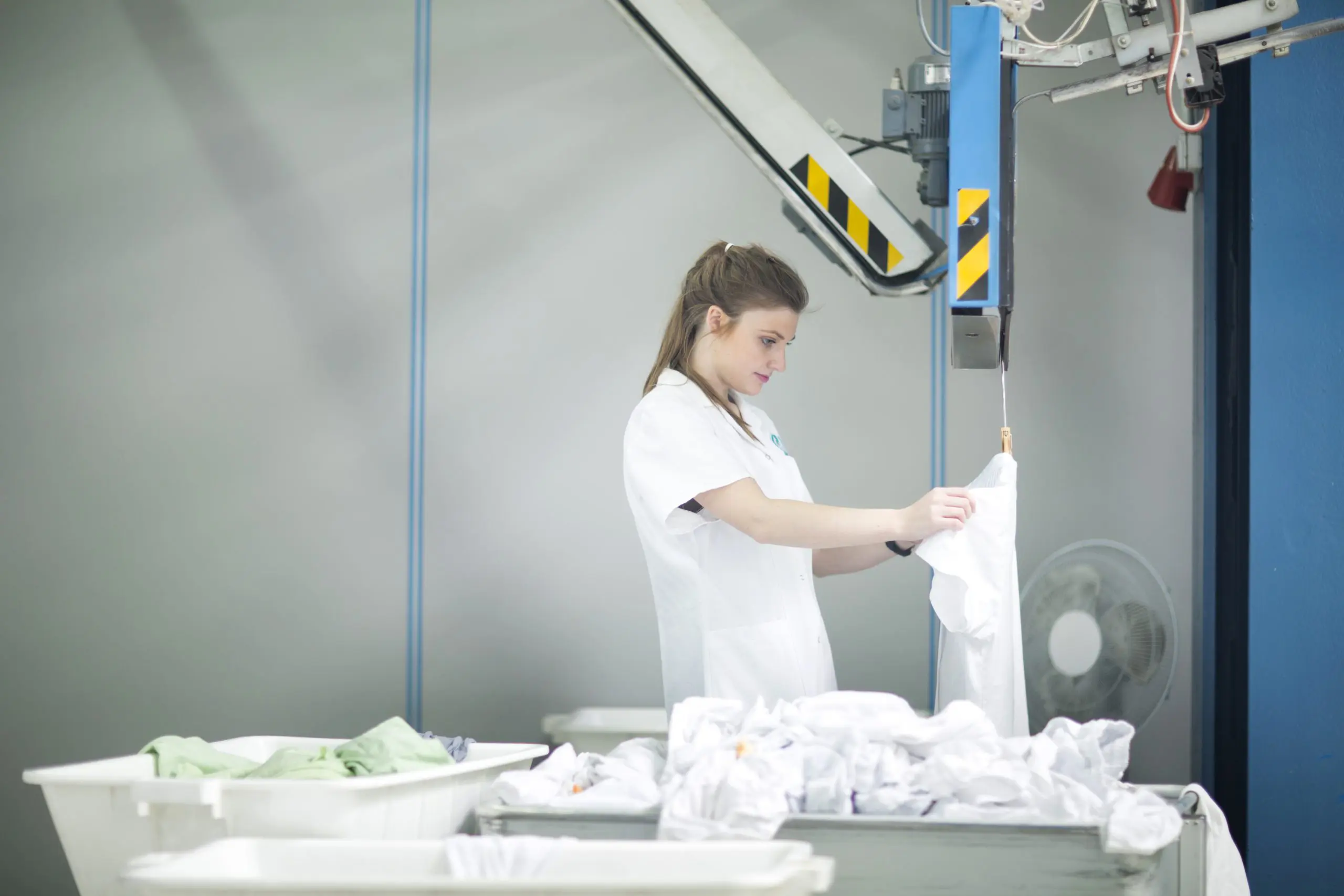 A woman in a white lab coat sorts white garments in a laundry facility. Multiple laundry bins filled with clothes are in front of her, and industrial laundry equipment is in the background.
