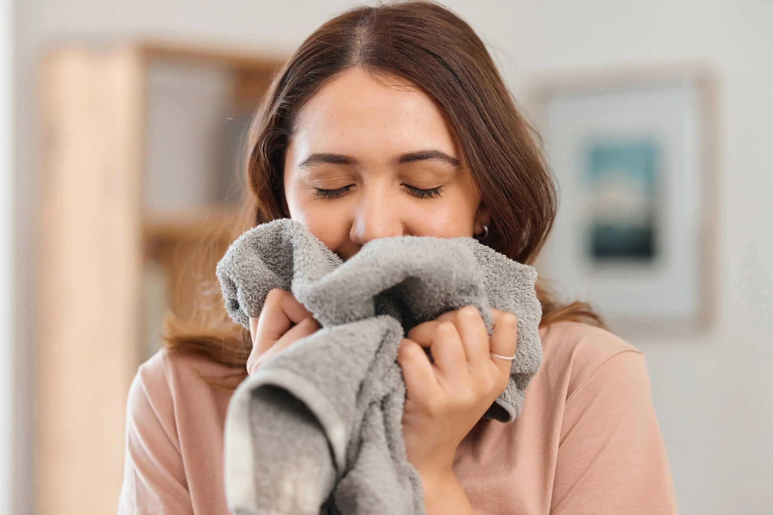 A woman with closed eyes holds a gray towel to her face, appearing to enjoy its texture or scent. She has brown hair and is indoors, with a slightly blurred background that includes a framed picture.