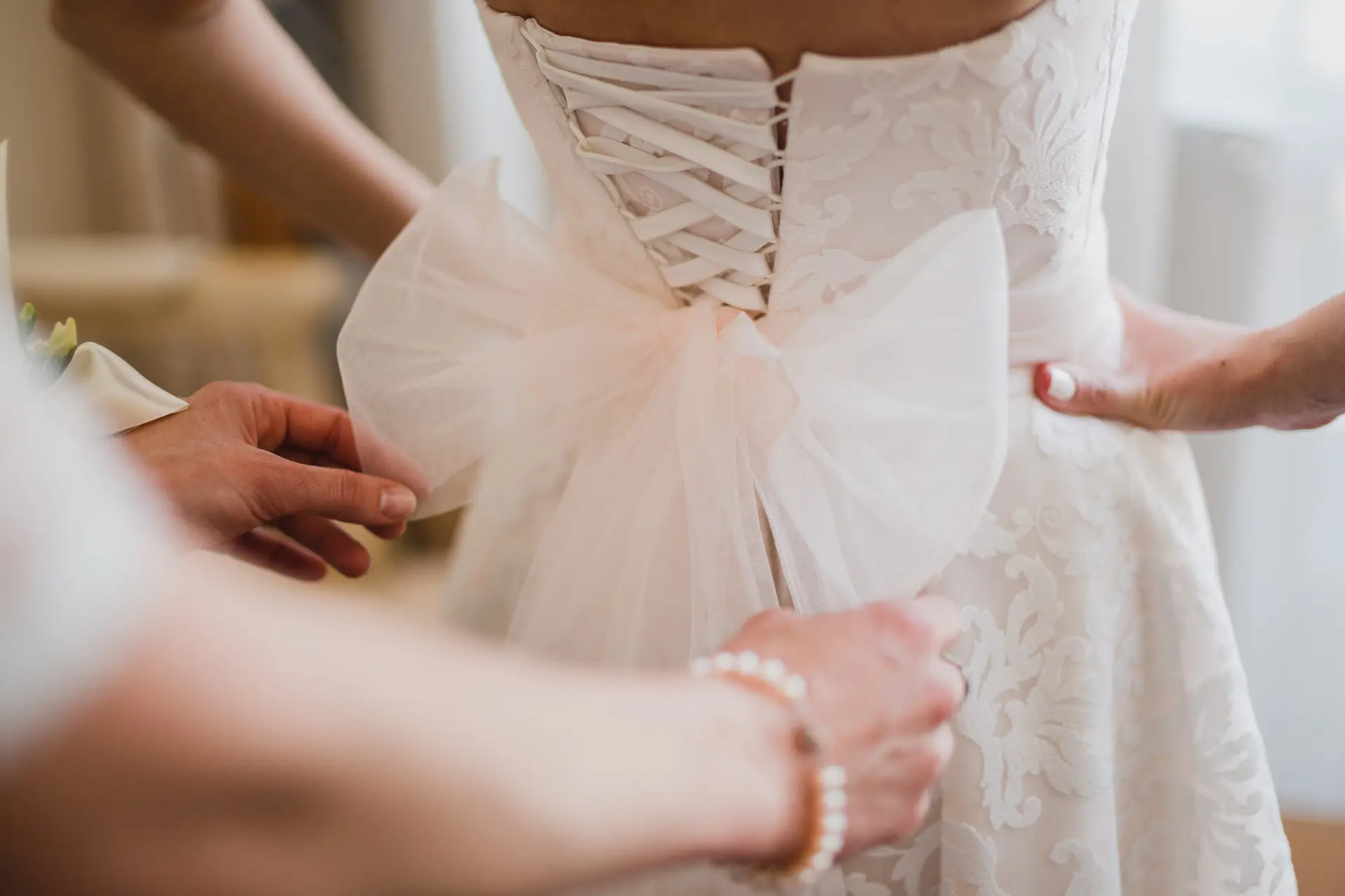 Close-up of a person adjusting the bow and lacing on the back of a white lace wedding dress worn by a bride.