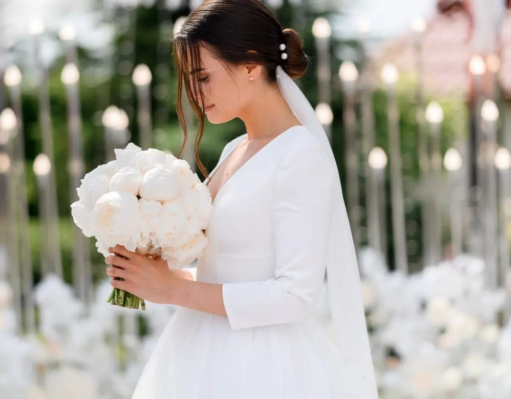 Bride in a white wedding dress and veil holding a bouquet of white flowers, surrounded by blurred greenery and decorative lights in the background.