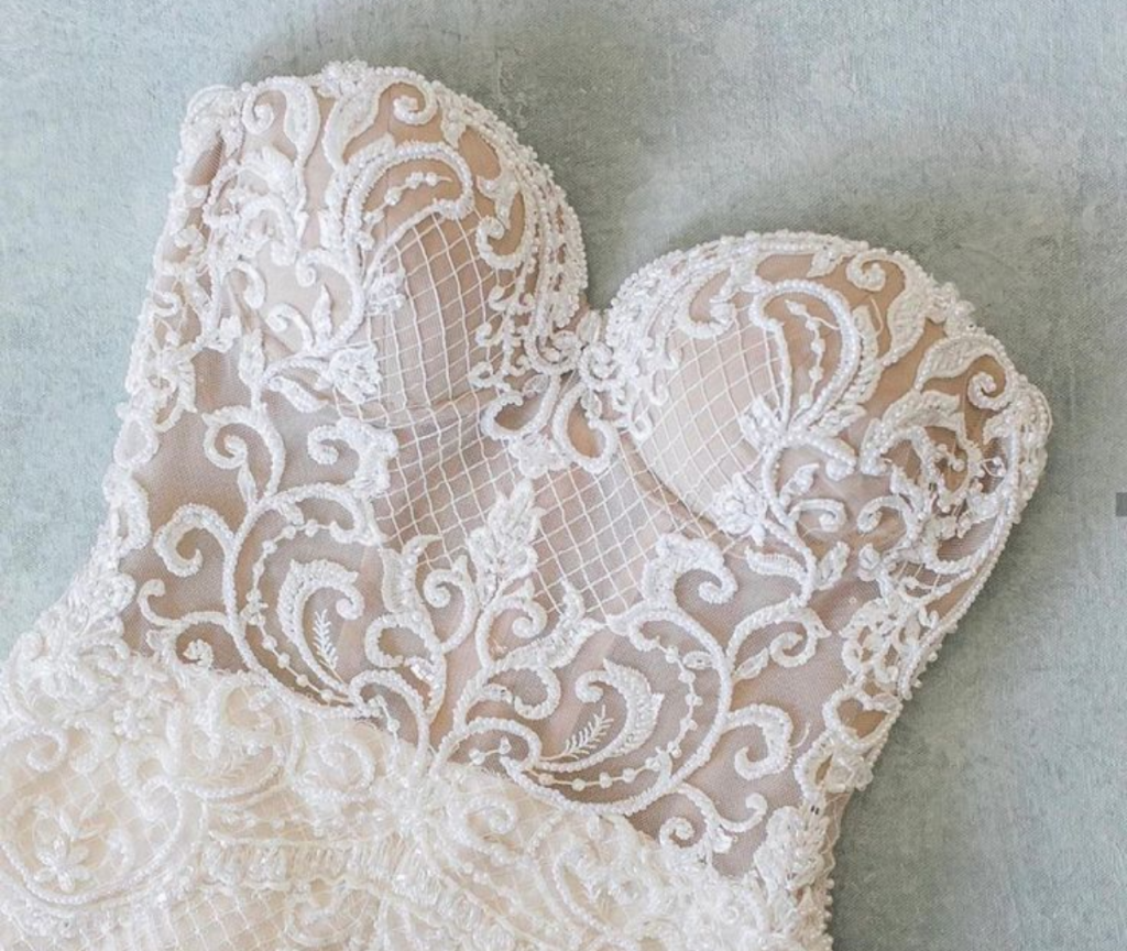 A close-up of a strapless wedding dress bodice featuring intricate lace embroidery and mesh detailing on a light background.