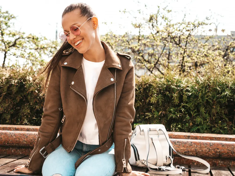 A young woman wearing a brown jacket and sunglasses sits on a bench outdoors, smiling, with a white bag beside her.
