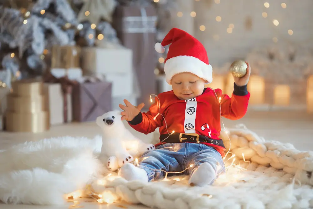 A baby dressed in a Santa outfit sits on a blanket surrounded by Christmas lights, holding an ornament, with a stuffed polar bear and a decorated tree in the background.