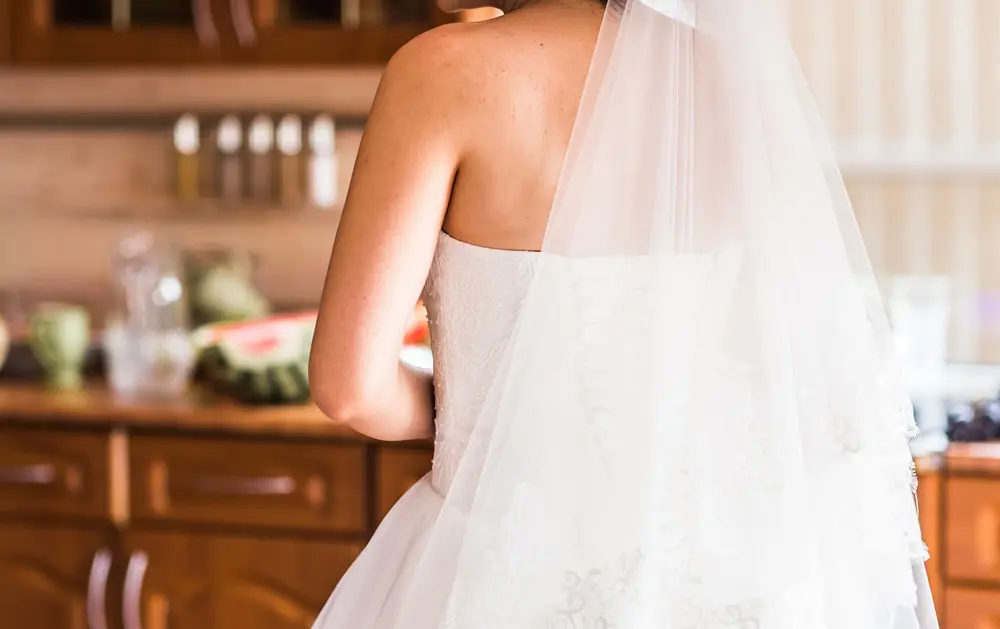 A bride in a white strapless wedding dress and veil stands indoors with her back to the camera, in a kitchen setting with blurred items on the counter.
