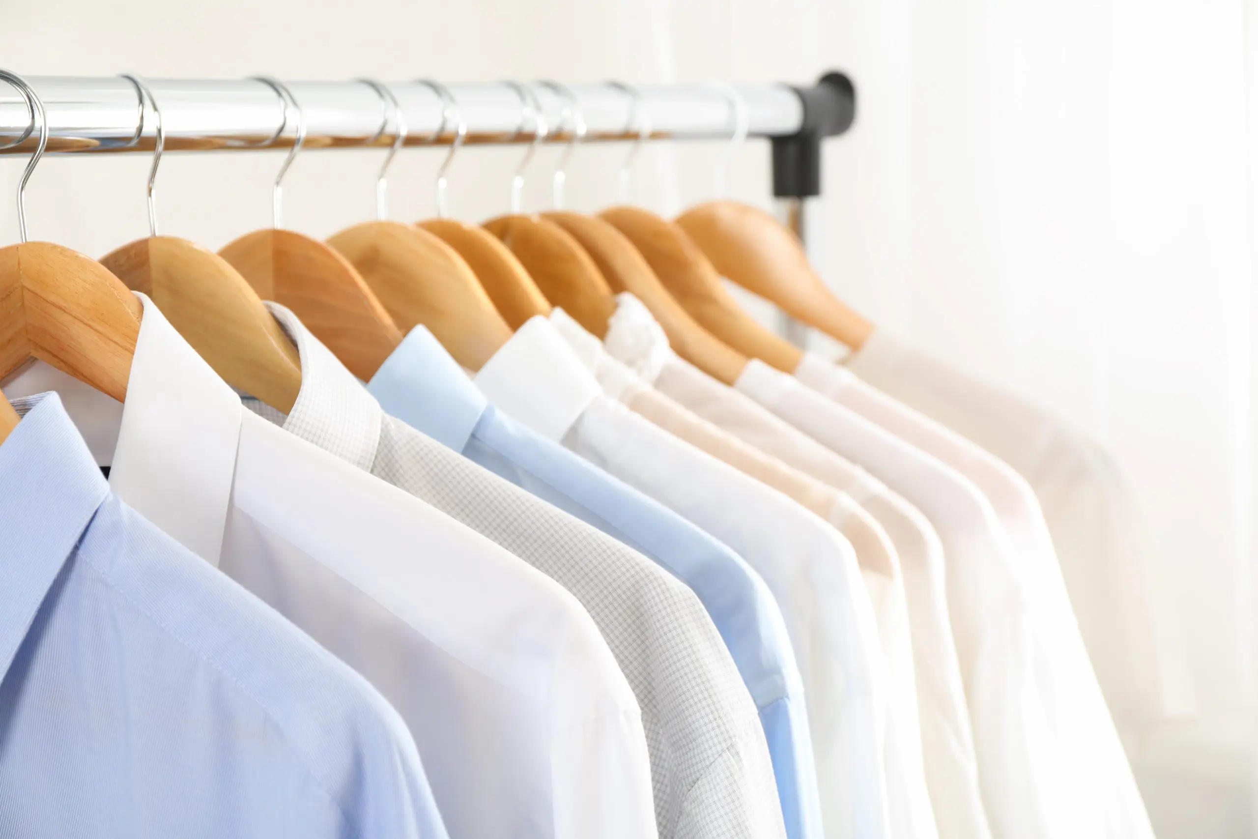 A row of neatly hung shirts in various shades of white and blue on wooden hangers, hanging from a metal rod.