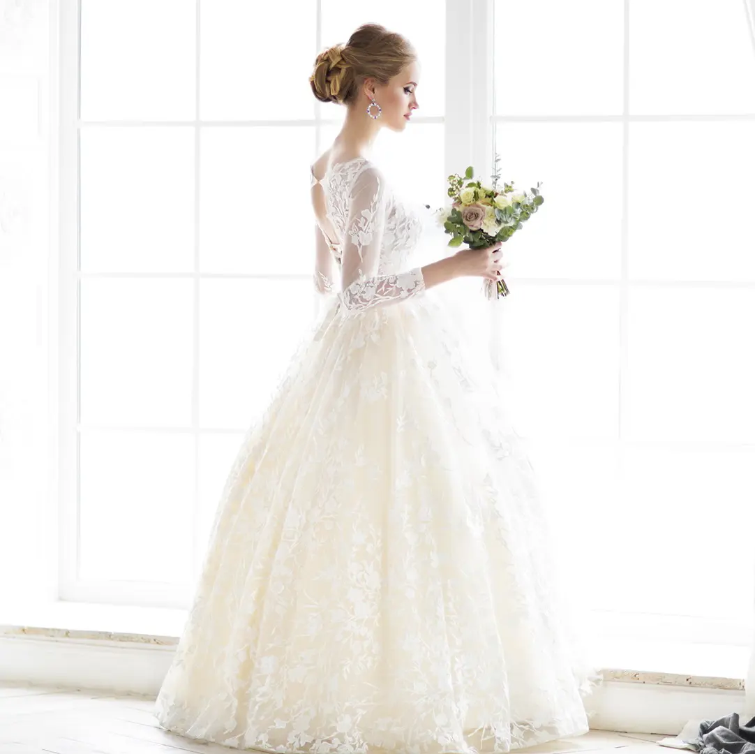 A bride in a white lace wedding dress stands by a large window holding a bouquet of flowers, looking to her right, blissfully unaware of the exceptional Dry Cleaning and Laundry Service in Marshall that helped make her gown pristine.
