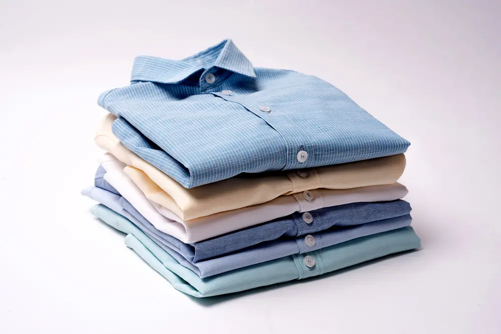 A neatly stacked pile of seven folded dress shirts in various pastel colors, including blue, beige, and white, against a plain background.