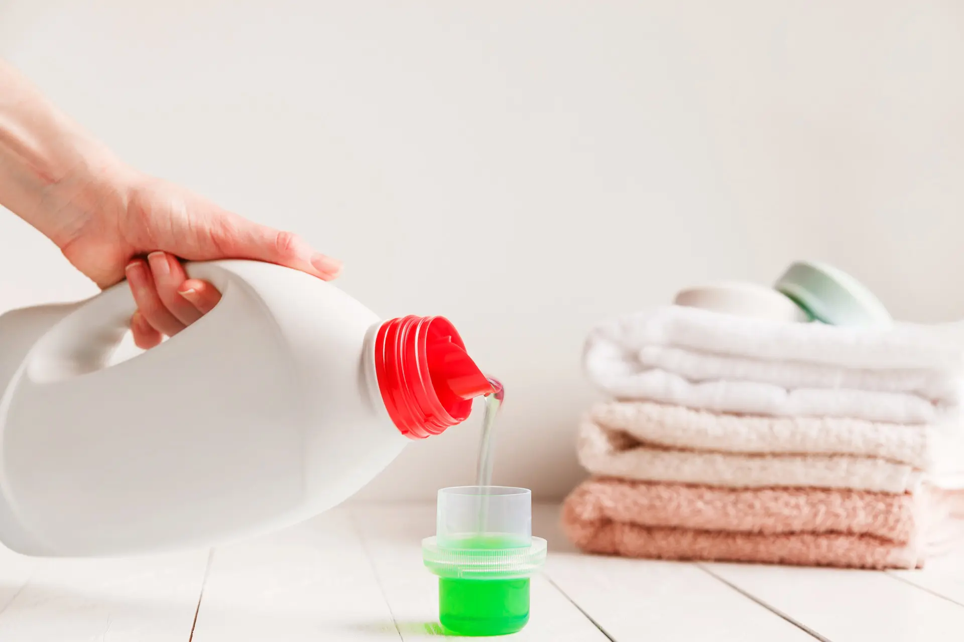 A person pours green liquid detergent into a cap near folded towels.