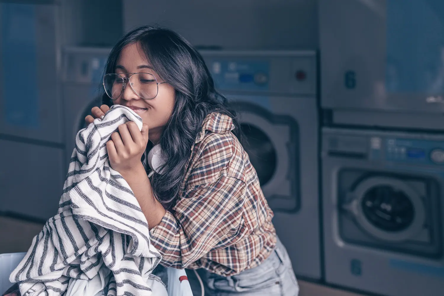 A woman in a plaid shirt and glasses enjoys the smell of a striped towel while holding a laundry basket in a laundromat.