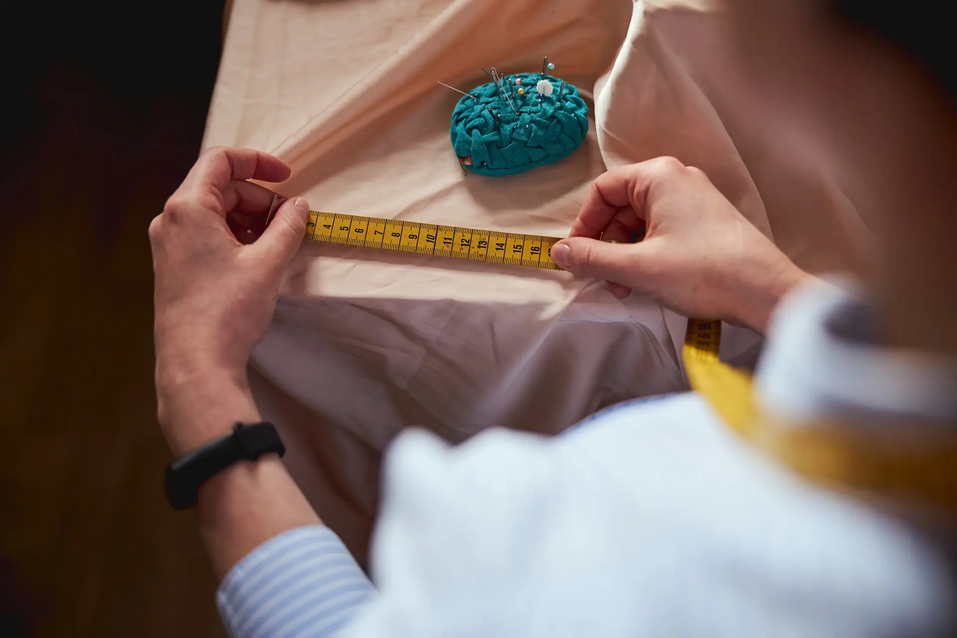 Person measuring beige fabric with a yellow tape measure; a blue pincushion with pins is on the table.