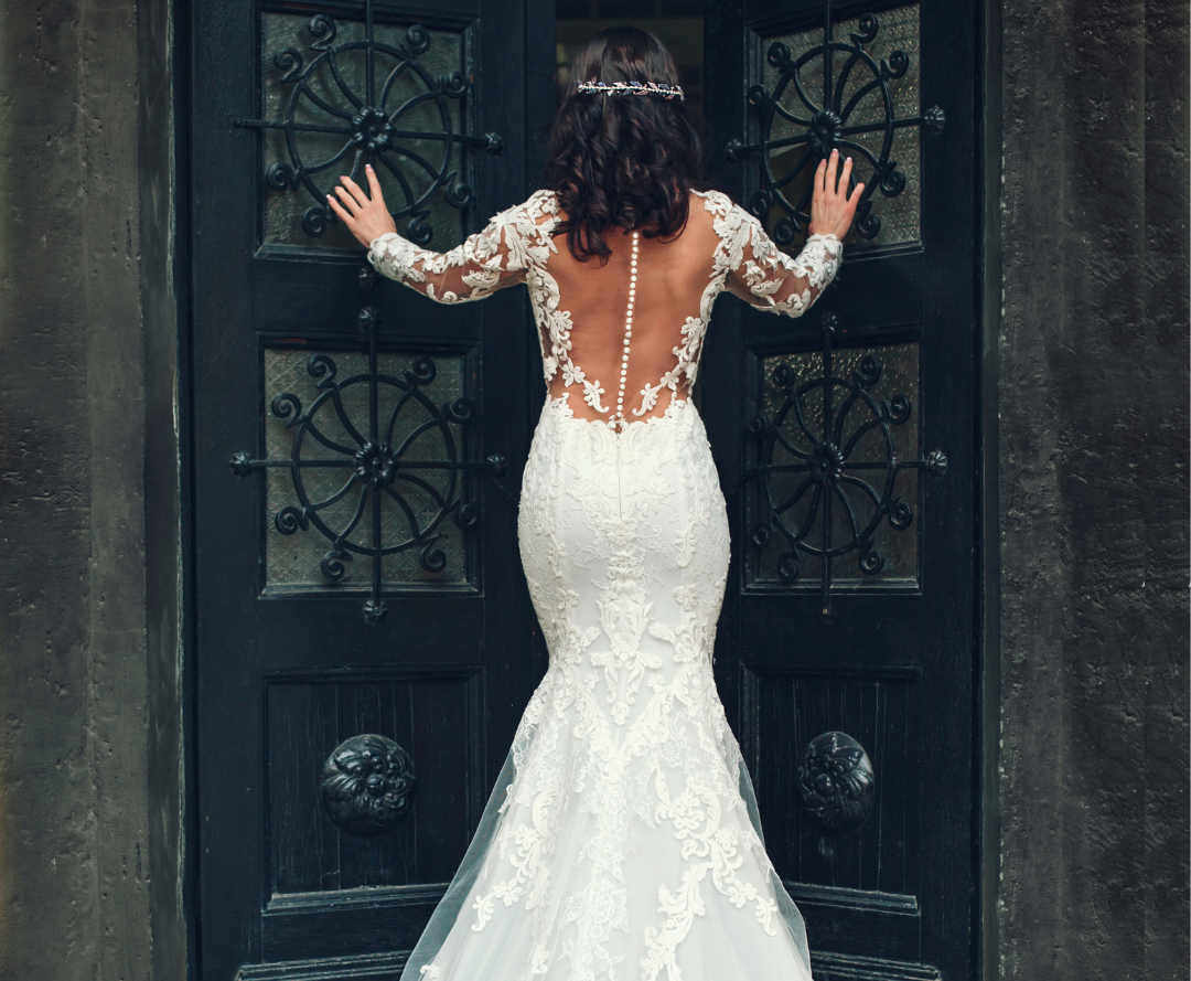 A woman in a lace-back wedding dress stands facing a large ornate black door with her hands on the door panels, her gown pristine as if cared for by expert cleaners for wedding gowns.