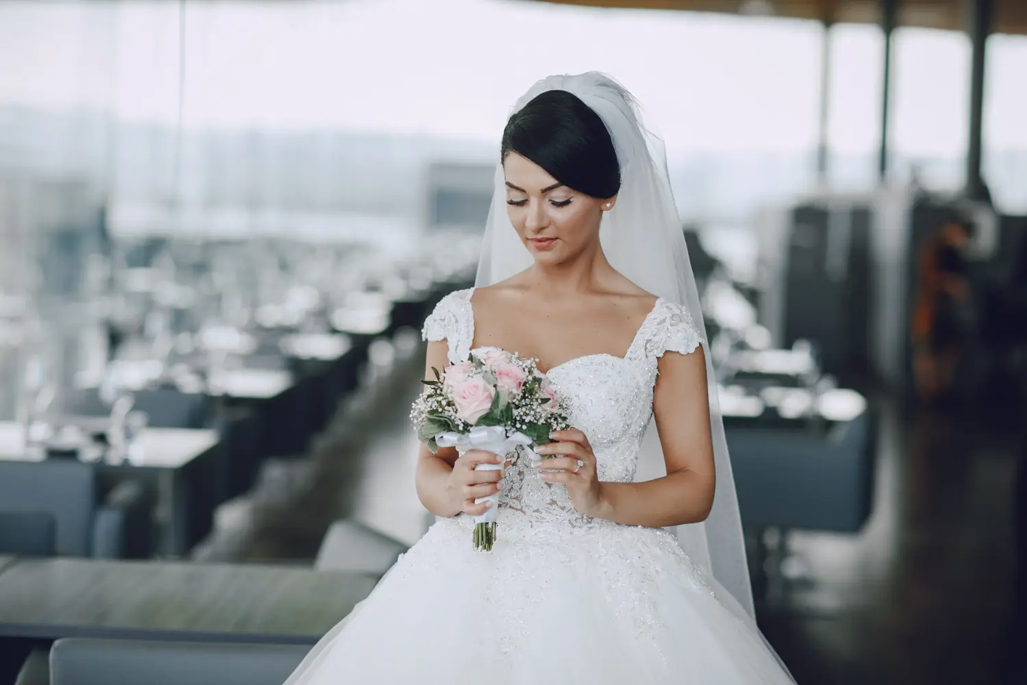 A bride in a white wedding dress and veil stands indoors, holding a bouquet of pink and white flowers, looking down at them.
