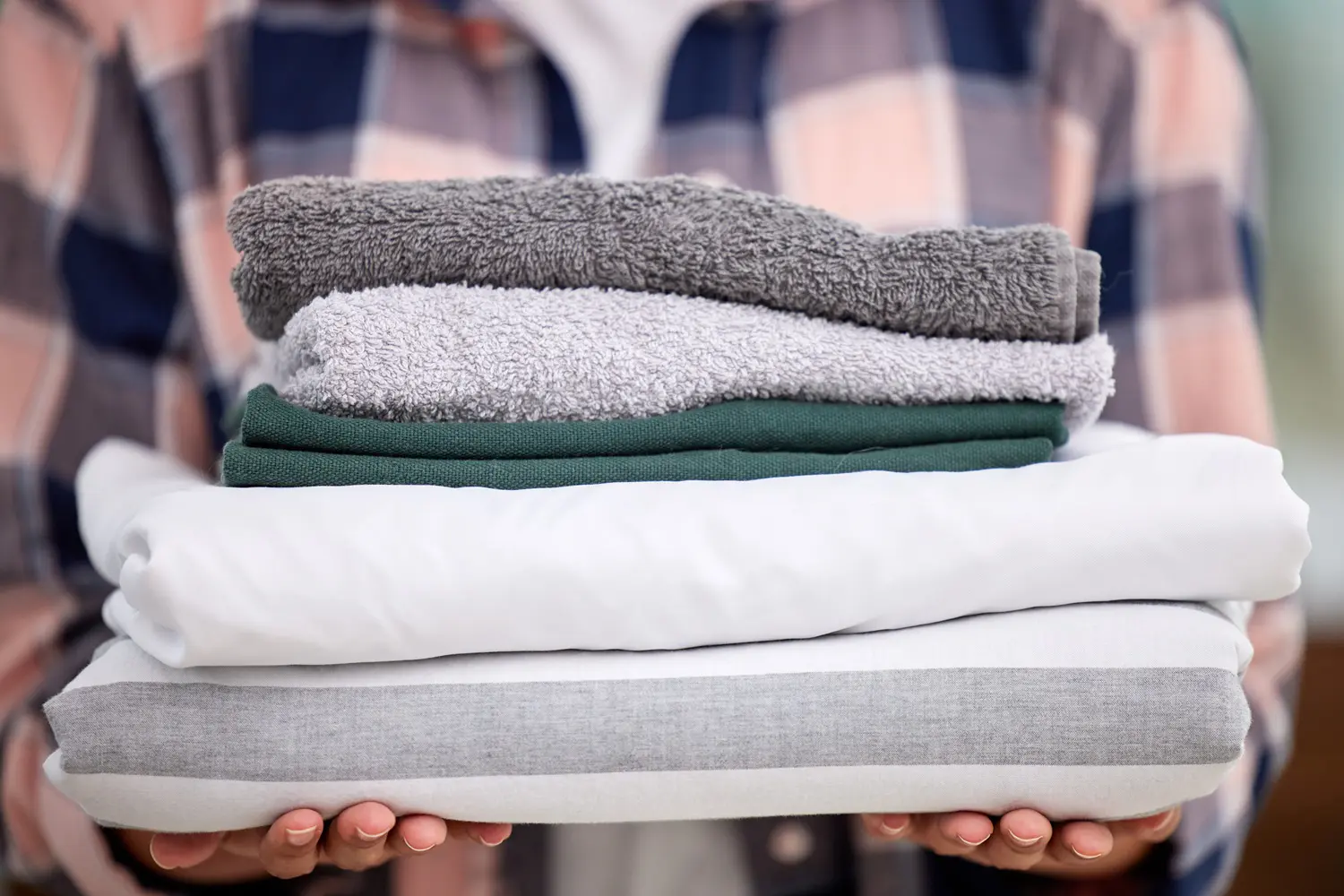 Person holding a stack of neatly folded towels and linens in various colors, including white, gray, and green, with a plaid shirt visible in the background.