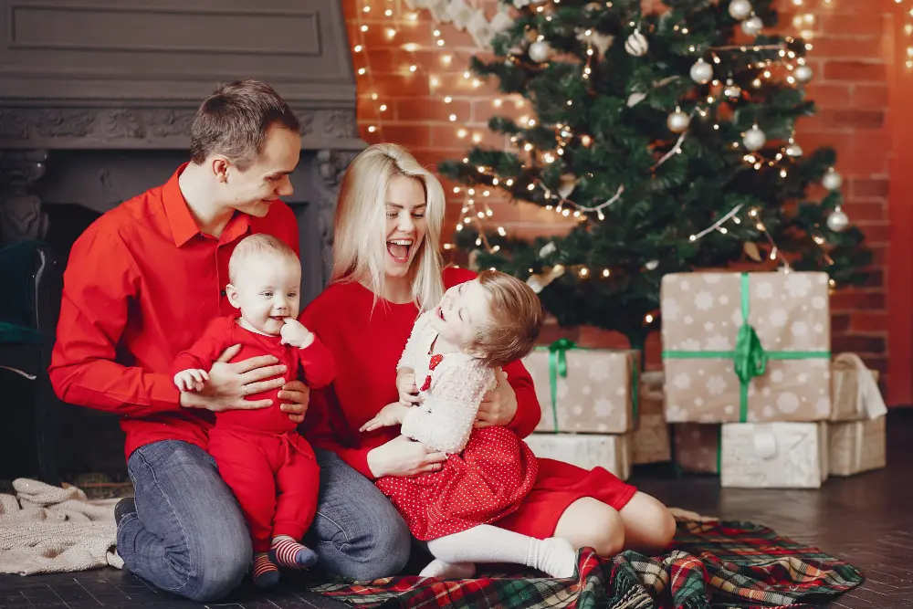 A family of four in red outfits sits on a blanket by a decorated Christmas tree and wrapped gifts, smiling and laughing together.