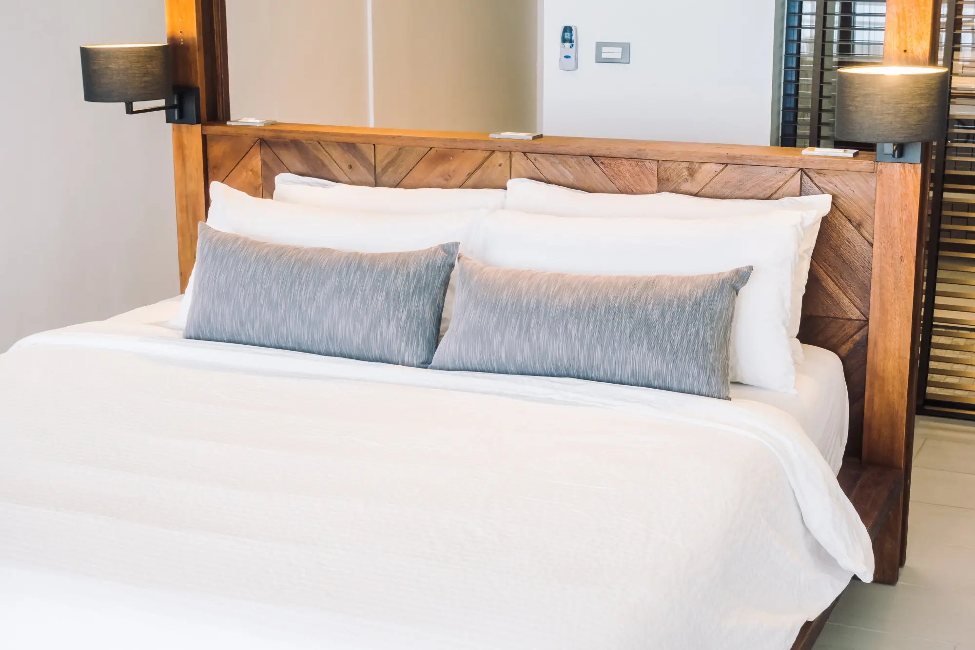 A neatly made bed with white bedding, two large gray pillows, and a wooden headboard with attached black wall lamps on each side.
