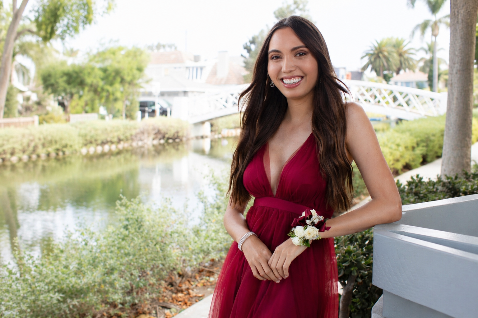 A woman in a sleeveless burgundy dress with a corsage stands by a canal, smiling at the camera; trees, a footbridge, and houses are in the background.