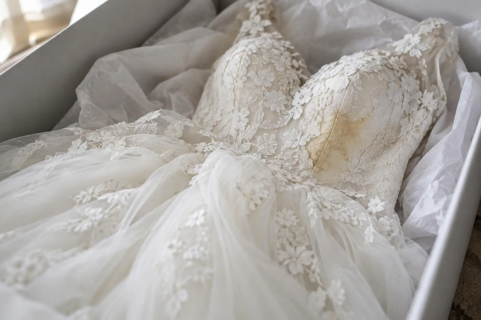 A white lace wedding dress with floral details is laid out in a box lined with tissue paper.