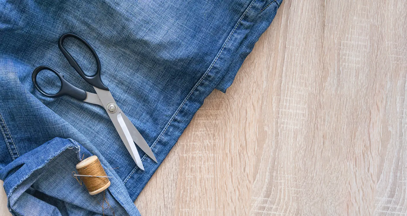 A pair of scissors, spool of thread, and denim fabric are arranged on a light wooden surface.
